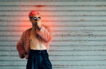 Giovane gay in occhiali da sole moderni e giacca di pelliccia elegante con capelli rosa contro muro grigio — Foto stock