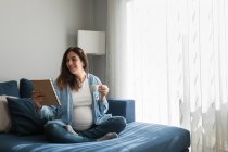 Позитивна вагітна жінка сидить на м'якому дивані з блокнотом і п'є гарячий напій — стокове фото