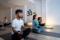 Afro-Américain avec un groupe de personnes diverses assis dans Lotus pose les yeux fermés et la médiation tout en pratiquant le yoga ensemble pendant les cours en studio — Photo de stock