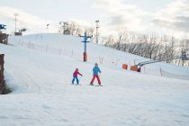 Full body faceless parent in warm sportswear and helmet teaching little kid to ski alongside snowy hill slope in winter ski resort — Stock Photo