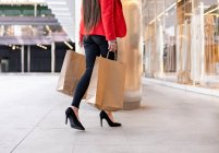 Crop comprador femenino con bolsas de papel y café para llevar de pie cerca de escaparate de la tienda en la ciudad - foto de stock