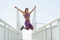 Atleta femenina satisfecha en ropa deportiva saltando con los brazos extendidos por encima del puente y mirando hacia otro lado durante el día - foto de stock
