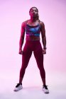 Молодая мускулистка афроамериканка в спортивной одежде с афрокосичками и руками на бедрах, смотрящая в неоновом свете — стоковое фото