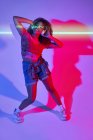 Mujer afroamericana feliz de cuerpo completo en pantalones cortos escuchando música favorita en auriculares y bailando con los ojos cerrados en luces de neón en el estudio - foto de stock