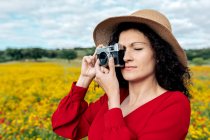 Femme souriante en chapeau prenant des photos sur caméra vintage sur prairie sous un ciel nuageux — Photo de stock