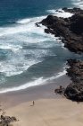 Desde arriba vista trasera de una viajera anónima admirando el océano espumoso desde la playa de arena entre montañas - foto de stock