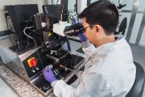 Vista lateral enfocada al hombre experto en guantes de bata de laboratorio que examina muestras a través de potentes lentes de microscopio mientras trabaja en un moderno laboratorio equipado - foto de stock