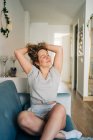 Positive attractive female in shorts sitting on cozy couch in living room stretching with arms up with eyes closed — Photo de stock