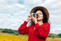 Mulher sorridente de chapéu tirando foto na câmera vintage no prado sob céu nublado — Fotografia de Stock