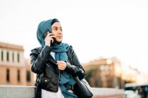 Mulher muçulmana alegre em lenço de cabeça tradicional em pé na rua da cidade e falando no telefone celular enquanto olha para longe — Fotografia de Stock