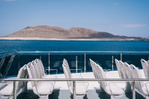 Пустые белые стулья на палубе круизного судна, плывущего в голубой морской воде с горой на берегу — стоковое фото