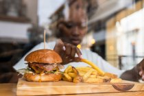 Through glass wall blurred African American female eating delicious fries and yummy hamburger served on wooden board on high table in fast food restaurant — Stock Photo