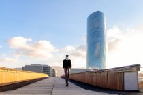 Unrecognizable ethnic male entrepreneur in sterile mask riding electric scooter on city bridge walkway against buildings under cloudy blue sky during pandemic — Stock Photo