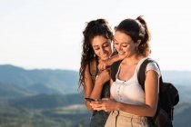 Счастливые молодые женщины-путешественницы в летней одежде, используя смартфон вместе, стоя на пышной солнечной холмистой местности — стоковое фото