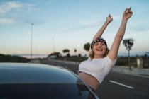 Молодая женщина в повседневной одежде и повязке с отпечатком американского флага, высунутая из окна машины и поднимая руки, наслаждаясь свободой во время путешествия на закате — стоковое фото