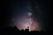 Baixo ângulo de silhueta de turista anônimo em pé no penhasco contra o céu estrelado brilhante à noite — Fotografia de Stock