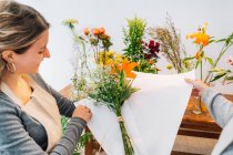Вид на урожай счастливая женщина-флористка в фартуке обертывая букет свежей апельсиновой лилии и цыганки в бумагу с анонимным коллегой — стоковое фото