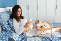 Весела вагітна жінка сидить на ліжку вранці і снідає під час перегляду смартфона — стокове фото