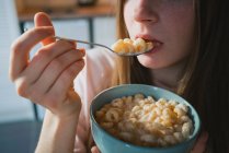 Ritaglia la giovane femmina con cucchiaio e ciotola godendo di gustosi anelli di mais mentre guarda la fotocamera in cucina — Foto stock