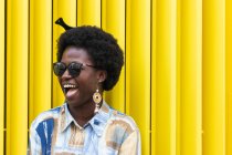 Portrait of young African American female with traditional comb on afro hair dressed in stylish colorful informal outfit smiling happily while standing against colorful yellow wall on urban street — Stock Photo