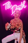 Attractive young bare shouldered female in white pants with bouquet of fresh colorful flowers sitting on stool against bright neon inscription in dark studio — Foto stock