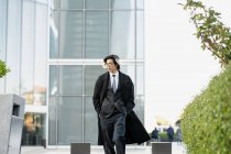 Auto-assegurada jovem empresário étnico masculino em terno formal e casaco andando com as mãos nos bolsos enquanto olha para longe na cidade — Fotografia de Stock