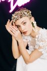 Charming tender young bride in white lace gown and luxurious floral wreath and earrings looking at camera against black background with neon lights — Photo de stock