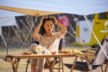 Hermosa mujer asiática étnica en gafas de sol sentada en la mesa mientras disfruta de un momento de relax en la zona de acampada durante las vacaciones mirando hacia otro lado - foto de stock