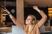 Mujer optimista sentada en el sofá y escuchando música en auriculares mientras disfruta de canciones con las manos levantadas - foto de stock