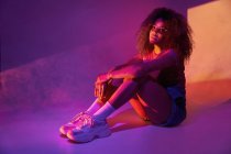 Full body young attractive African American female in shorts and trendy sunglasses sitting on floor in neon lights and embracing knees while looking at camera — Stock Photo