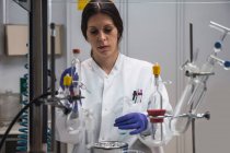 Концентрированная женщина-ученый в белом халате и перчатках проводит химический эксперимент с веществом и шприцем во время работы в современной лаборатории — стоковое фото