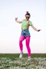 Corpo inteiro encantado jovem mulher vestindo shorts e calças rosa brilhante saltando alegremente com o braço levantado na exuberante clareira gramada na natureza — Fotografia de Stock