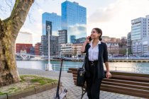 Lächelnde ethnische Unternehmerin lehnt auf Holzbank und telefoniert, während sie auf Stadtbank gegen Roller wegschaut — Stockfoto