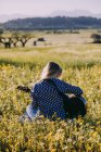 Vue arrière d'une femme hipster méconnaissable réfléchie assise sur une prairie à la campagne jouant de la guitare pendant la lumière du soleil d'été — Photo de stock