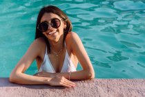Joven alegre turista étnica femenina en trajes de baño y accesorios apoyados con las manos en la piscina a la luz del sol - foto de stock