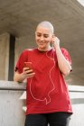 Joven mujer homosexual en camiseta y auriculares con teléfono celular mirando a la pantalla mientras escucha música - foto de stock