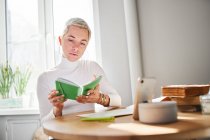 Une astrologue adulte lit un bloc-notes au bureau avec un smartphone et des livres à la maison au soleil — Photo de stock