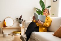 Зміст середнього віку жінка розслабляється на дивані і читає цікаву історію, насолоджуючись вихідними вдома — стокове фото