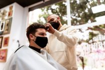 Male hairstylist in eyeglasses making haircut to adult client in hairdressing salon during COVID 19 pandemic — Photo de stock