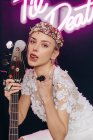 Energetic rebellious young woman in elegant white bridal dress and wreath with guitar in hand making horn gesture and sticking tongue out in studio with neon inscription — Stock Photo