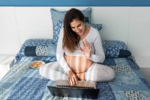 Dall'alto di una donna incinta sorridente che parla in videochiamata su netbook e agita la mano mentre siede su un comodo letto a casa — Foto stock