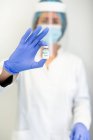 Female doctor in latex gloves and face shield standing with chemical liquid in glass vial preparing to vaccinate patient in clinic during coronavirus outbreak — Stock Photo