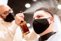 Male stylist in textile mask spraying haircut of client with liquid from bottle in barbershop — Foto stock