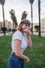 Vue latérale de la jeune femme en tenue décontractée et bandeau avec drapeau américain couvrant la bouche et riant tout en se tenant debout dans la rue urbaine — Photo de stock