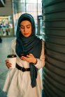 Etnia feminina no hijab em pé na estação e navegando telefone celular enquanto espera pelo trem — Fotografia de Stock