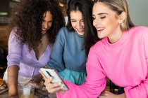 Smiling young female friends wearing casual clothes browsing mobile phones while gathering for lunch in restaurant — Stock Photo