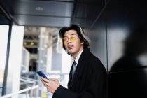 Молодой внимательный азиатский мужчина исполнительный в формальной одежде и солнцезащитных очках текстовые сообщения о глядя в сторону — стоковое фото