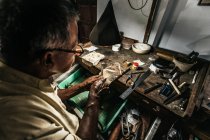 From above of concentrated ethnic elderly male jeweler in casual clothes and eyeglasses sharpening instruments while working at wooden table in traditional workshop — Stock Photo
