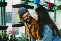 Conteúdo casal elegante abraçando em estufa enquanto está perto da prateleira e colhendo flor em vaso florescendo — Fotografia de Stock