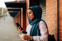Vista lateral da fêmea étnica no hijab em pé na plataforma na estação ferroviária e navegando telefone celular enquanto espera pelo trem — Fotografia de Stock